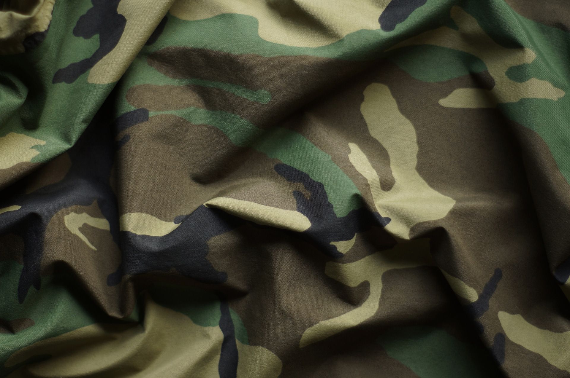 Camouflage fabric with green, brown, and black splotches, crumpled with visible folds.