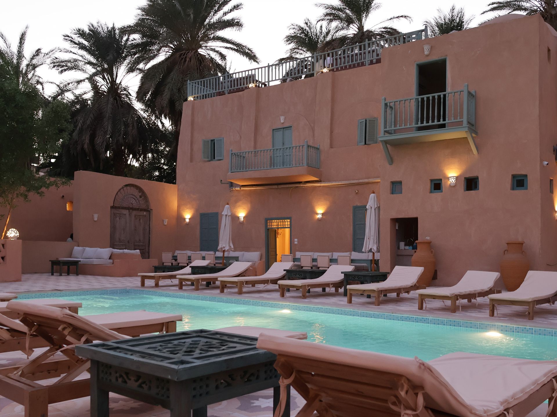 Poolside view of a peach-colored building with light blue accents, lounge chairs, and palm trees.