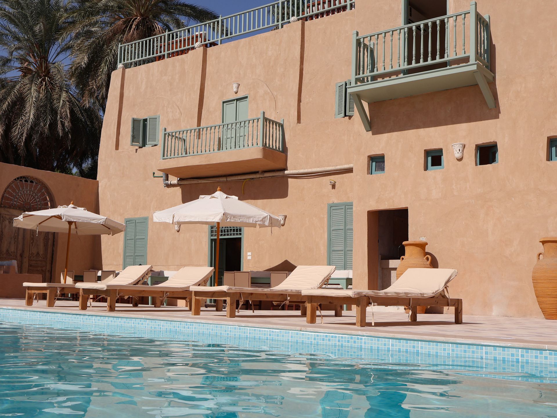 Poolside view of a peach-colored building with balconies and light green shutters. Lounge chairs and umbrellas.
