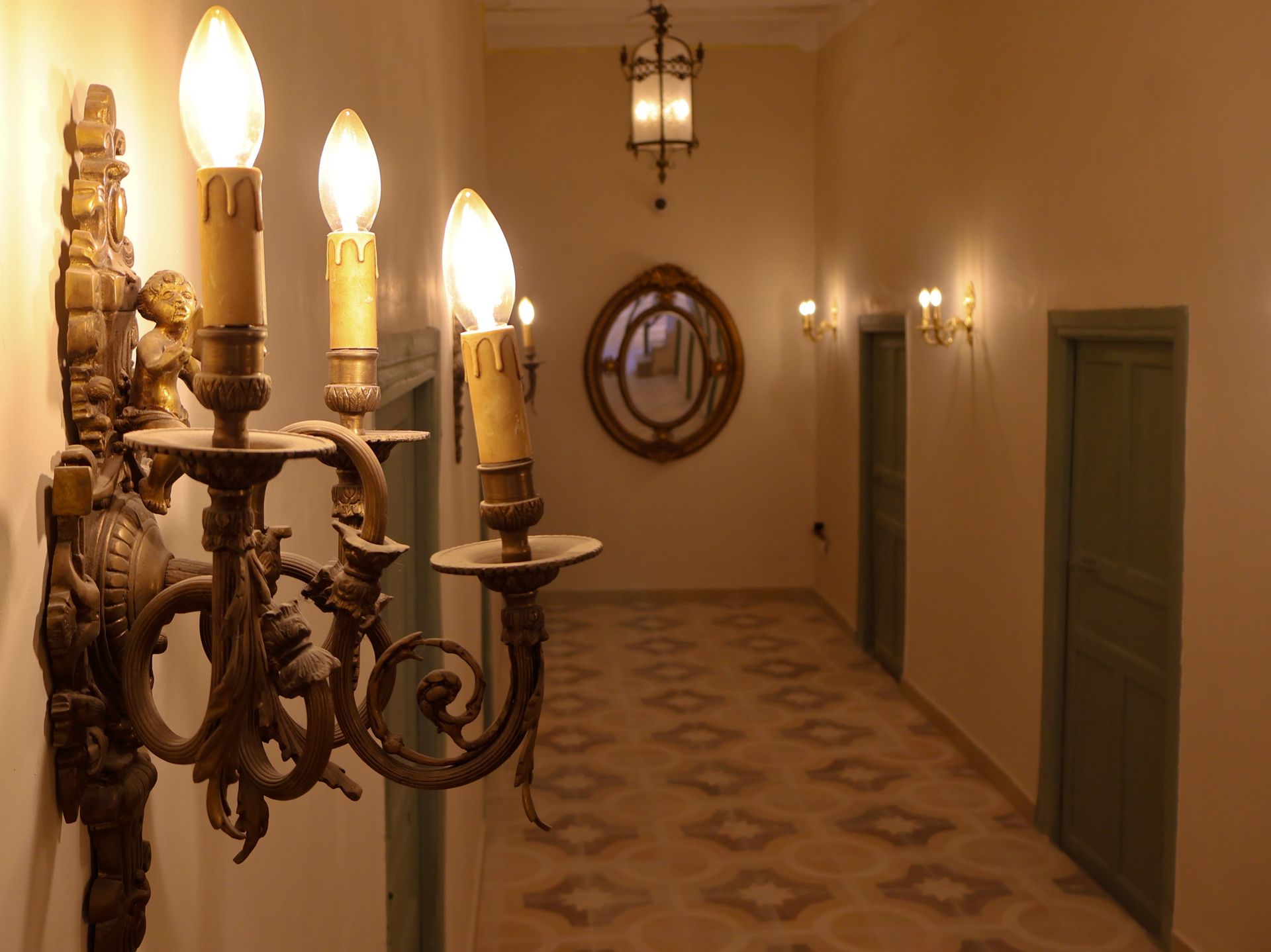 Ornate sconce with lit bulbs in a hallway with decorative flooring, a mirror, and closed green doors.