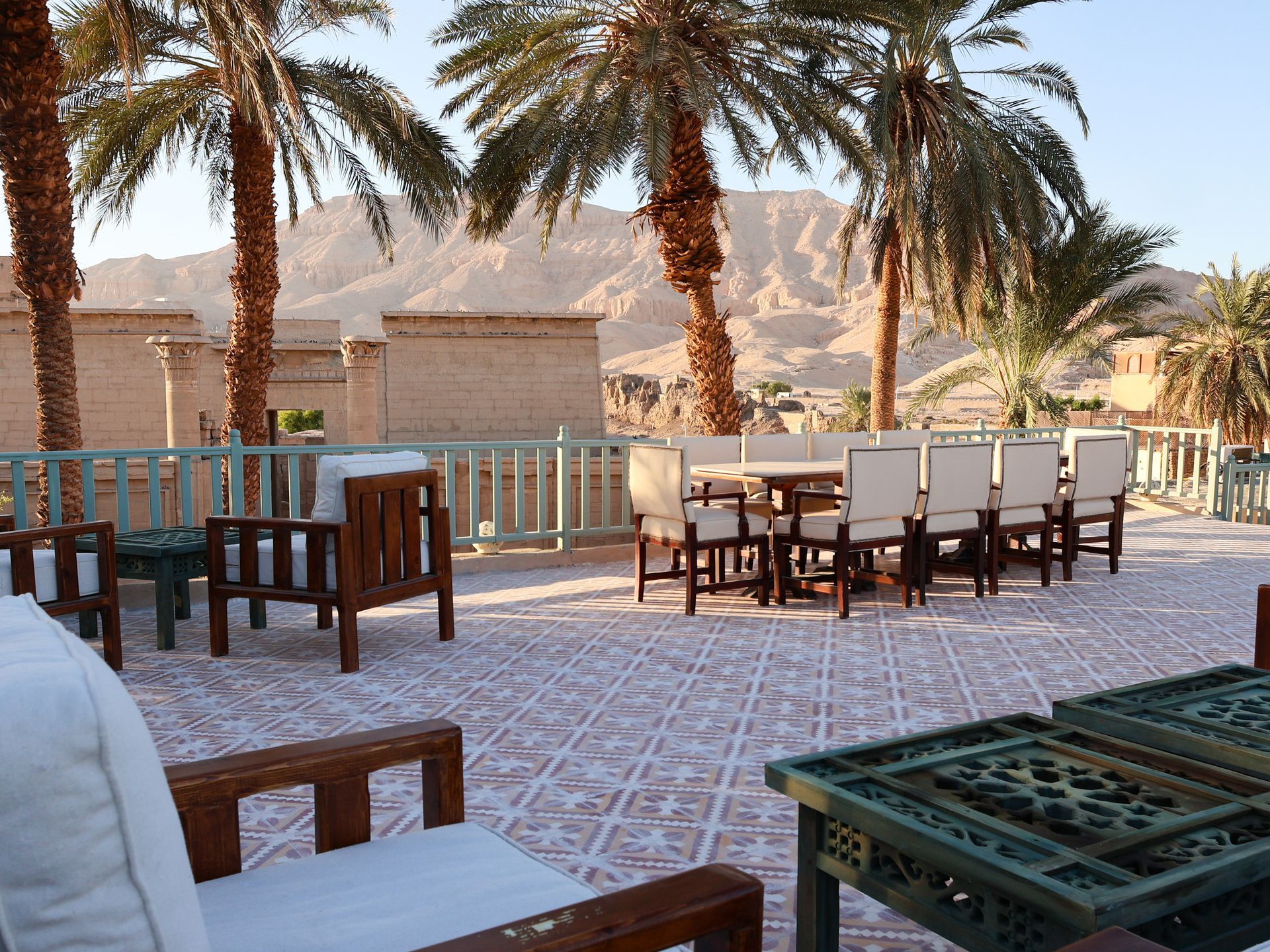 Outdoor patio with dining tables and chairs, palm trees, and a mountain backdrop.