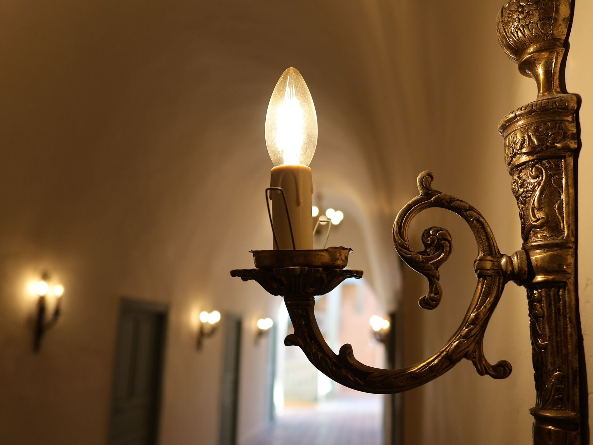 Close-up of a lit candle-shaped bulb in a brass sconce, with a dimly lit hallway in the background.