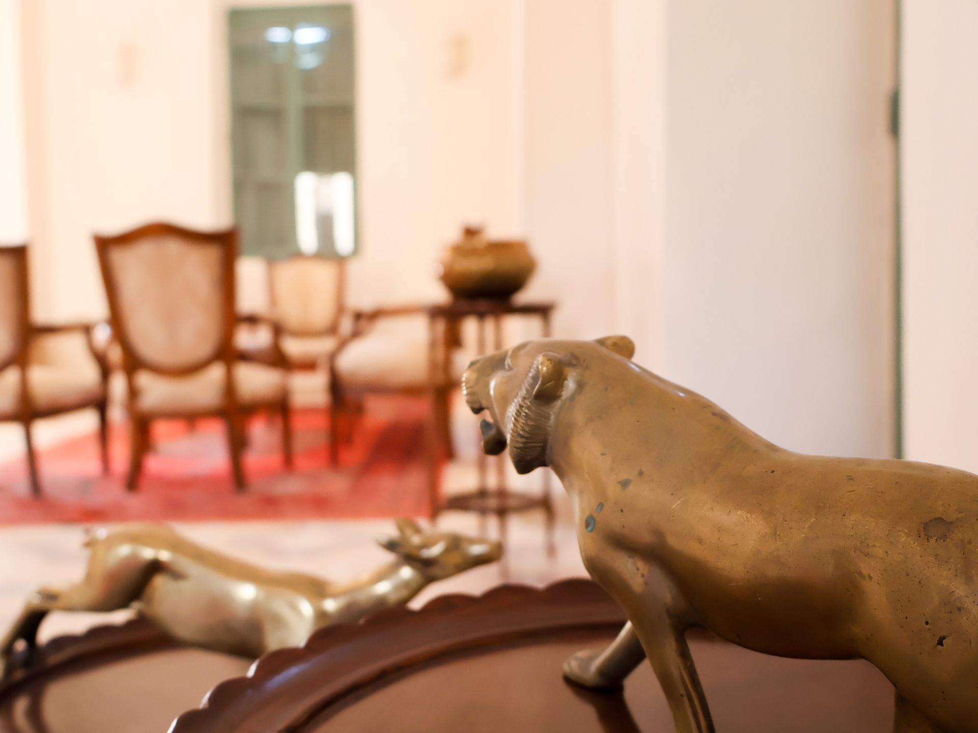 Brass lion and gazelle sculptures on a dark table, with chairs and a vase in a blurred background.