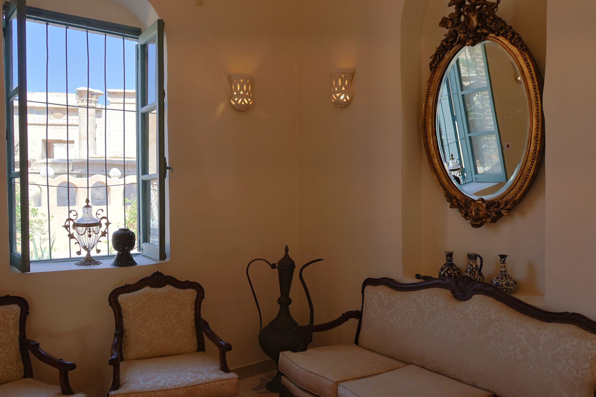 Cozy room with ornate mirror, sofa, chairs, and window with view. Light walls and fixtures.