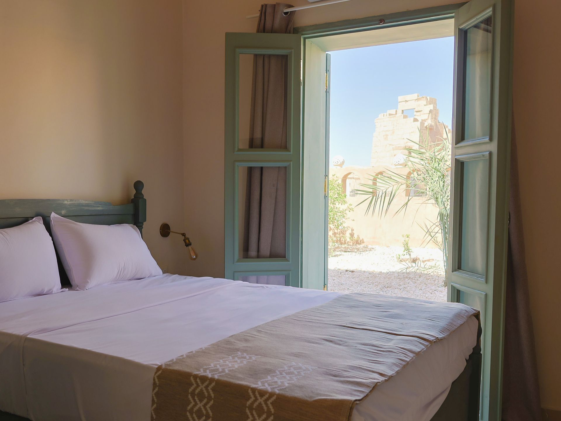 Bedroom with open teal-framed doors to a courtyard. White linens on a bed, beige walls, sunny exterior.