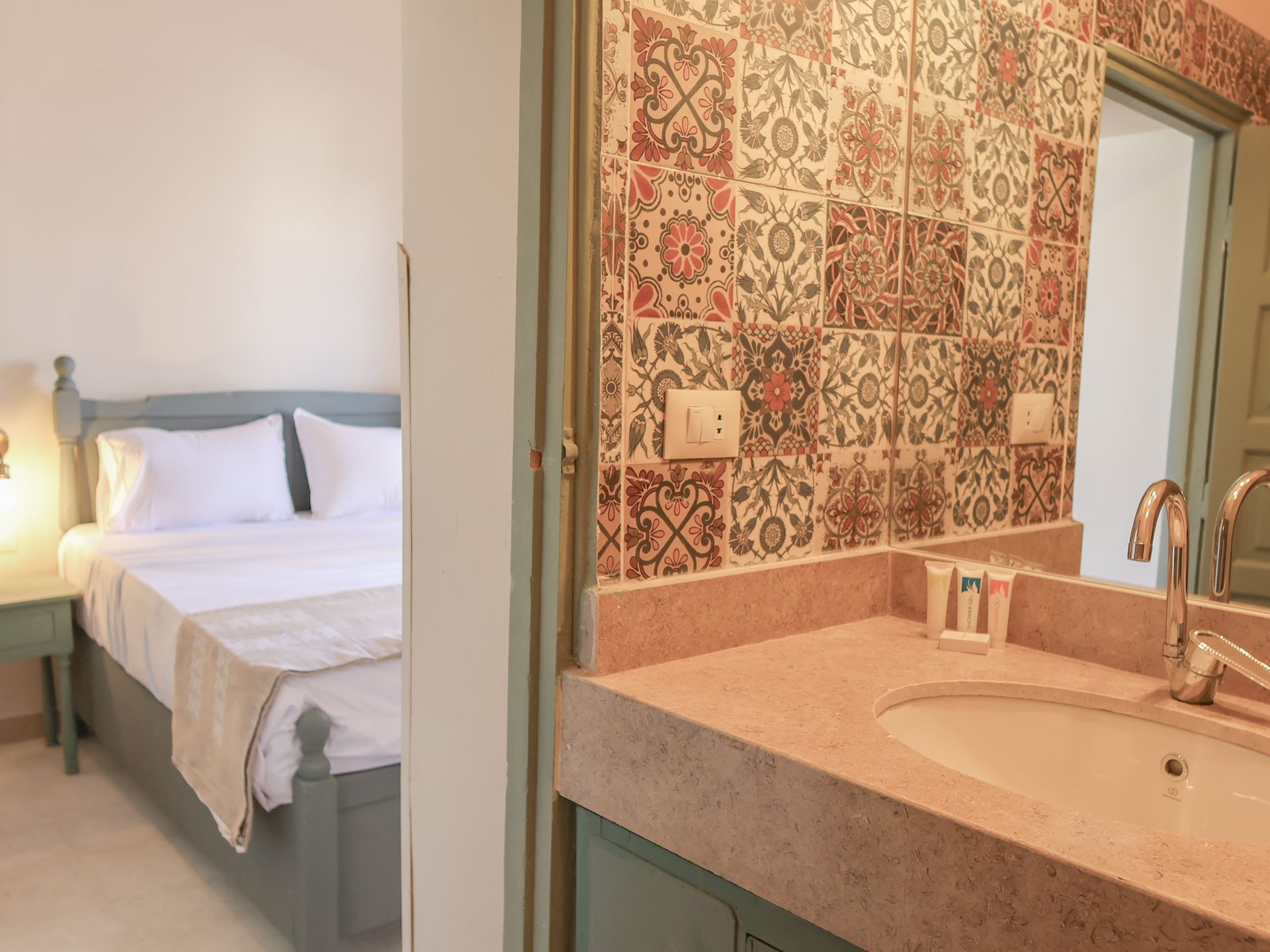 Bathroom with floral tile backsplash and view of a bedroom with a bed and bedside table.