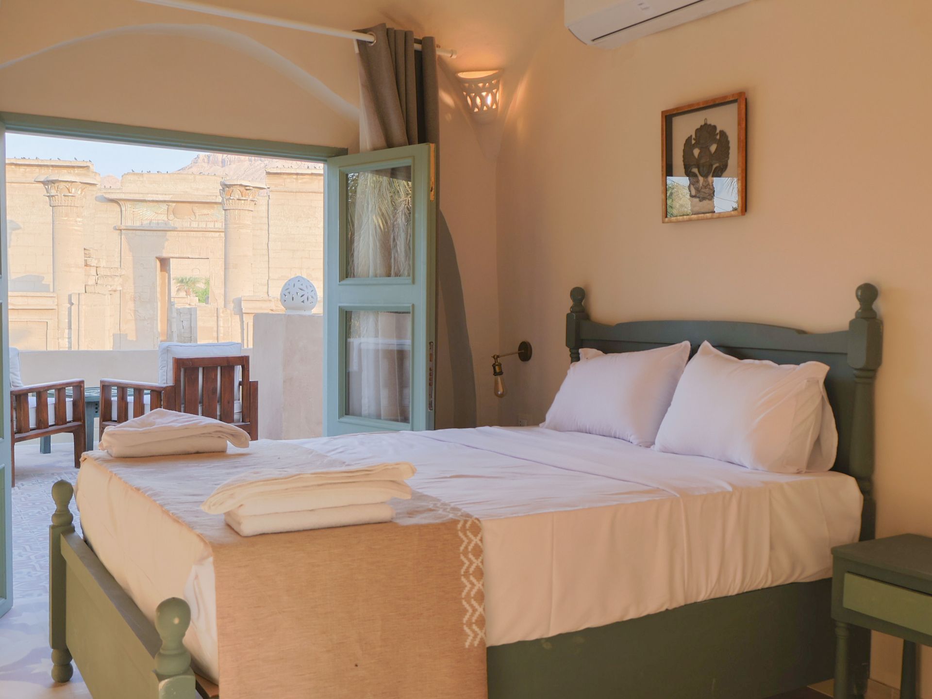 Bedroom with a bed and balcony overlooking an ancient structure; green accents, natural light.