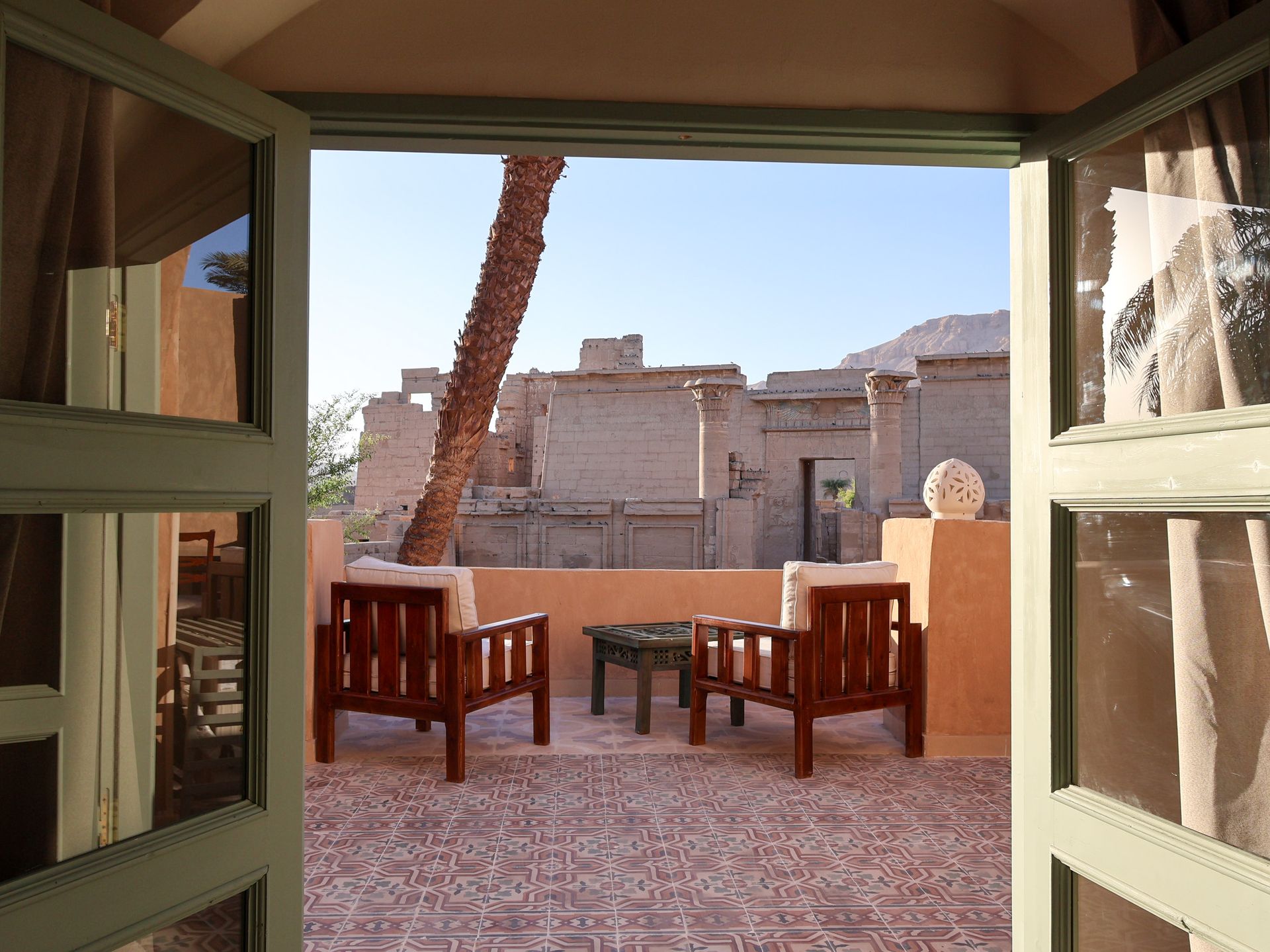 Two wooden chairs and table on a patio, viewed through open green-framed doors; ancient ruins and a palm tree in the background.