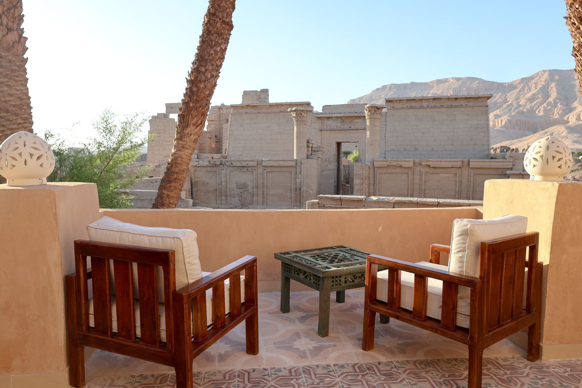 Two wooden chairs and table on a patio, overlooking ancient stone structures and mountains.