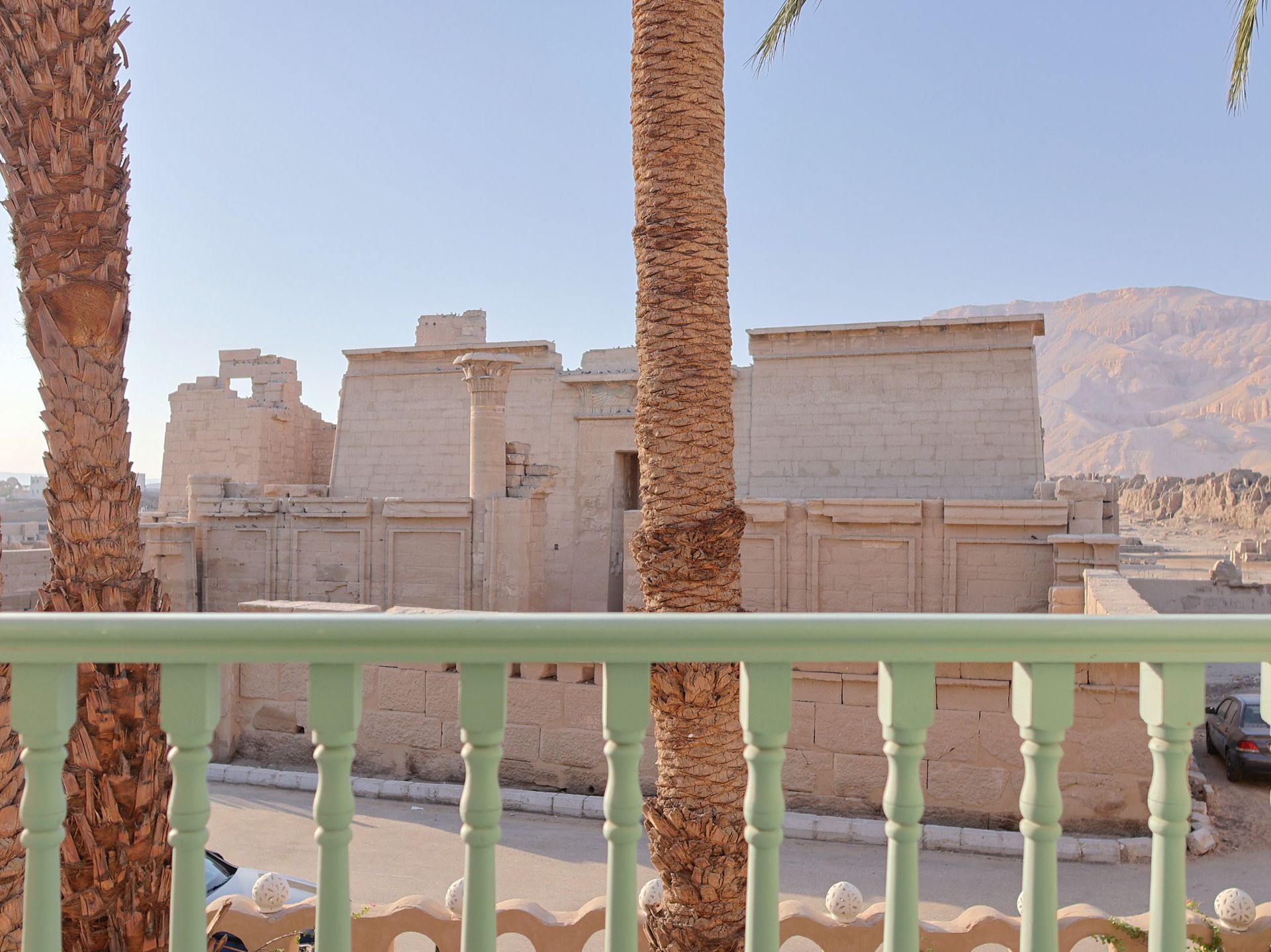 Ancient stone temple seen through a green railing and palm trees, with mountains in the background under a clear sky.