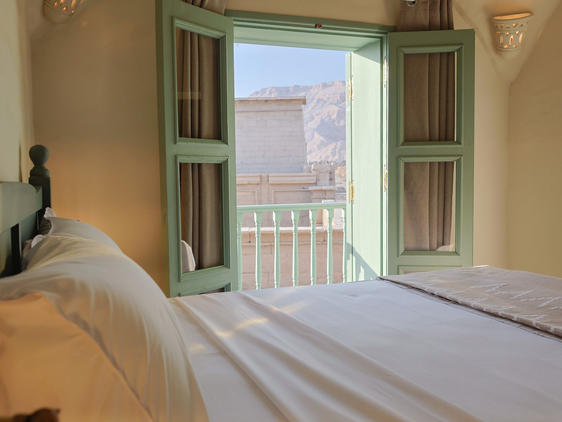 Bedroom with open, turquoise window frames overlooking a balcony. Soft bed with white linens.
