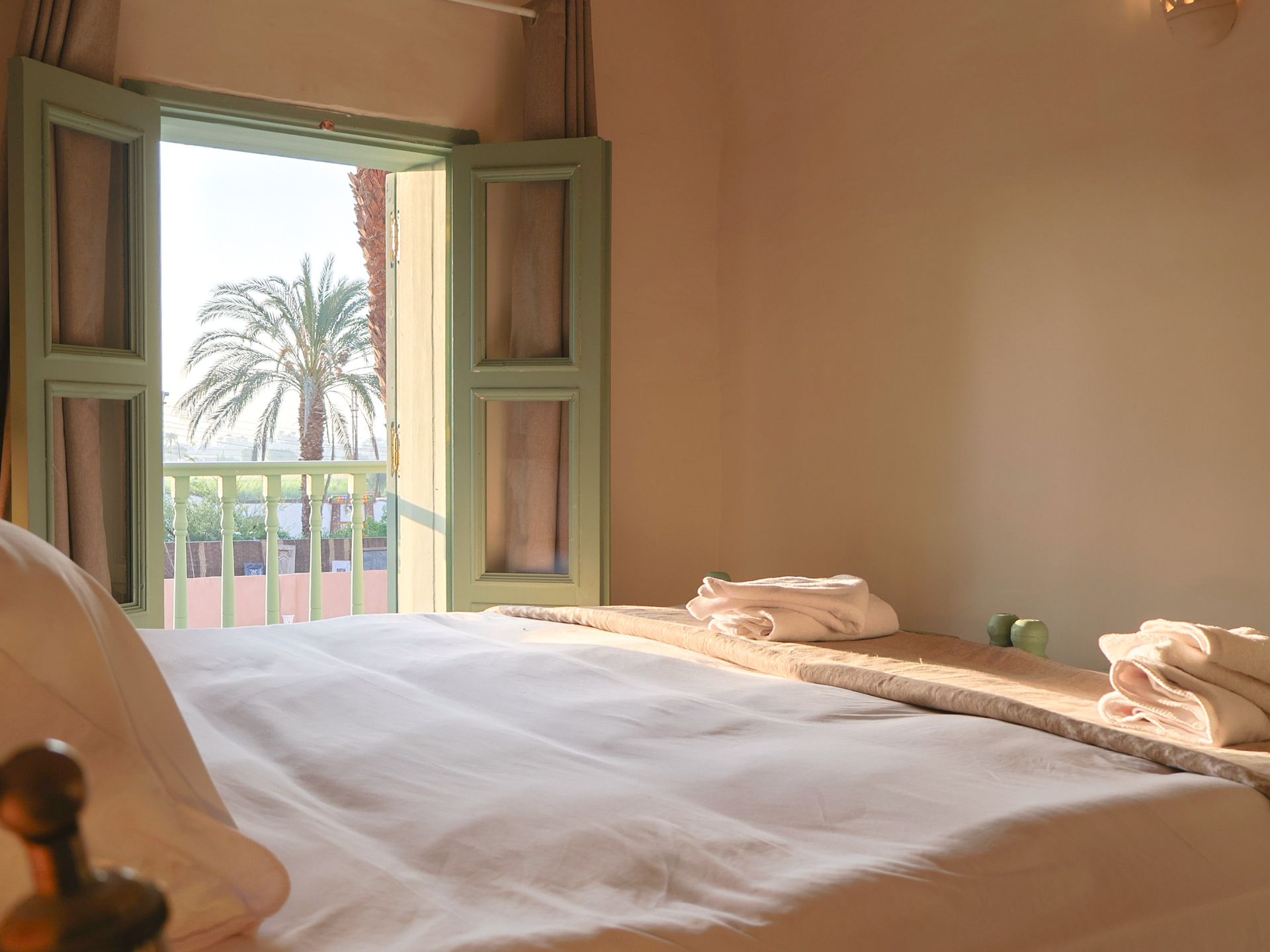 Bedroom with open window overlooking palm tree. White bedding, green shutters, sunlit scene.