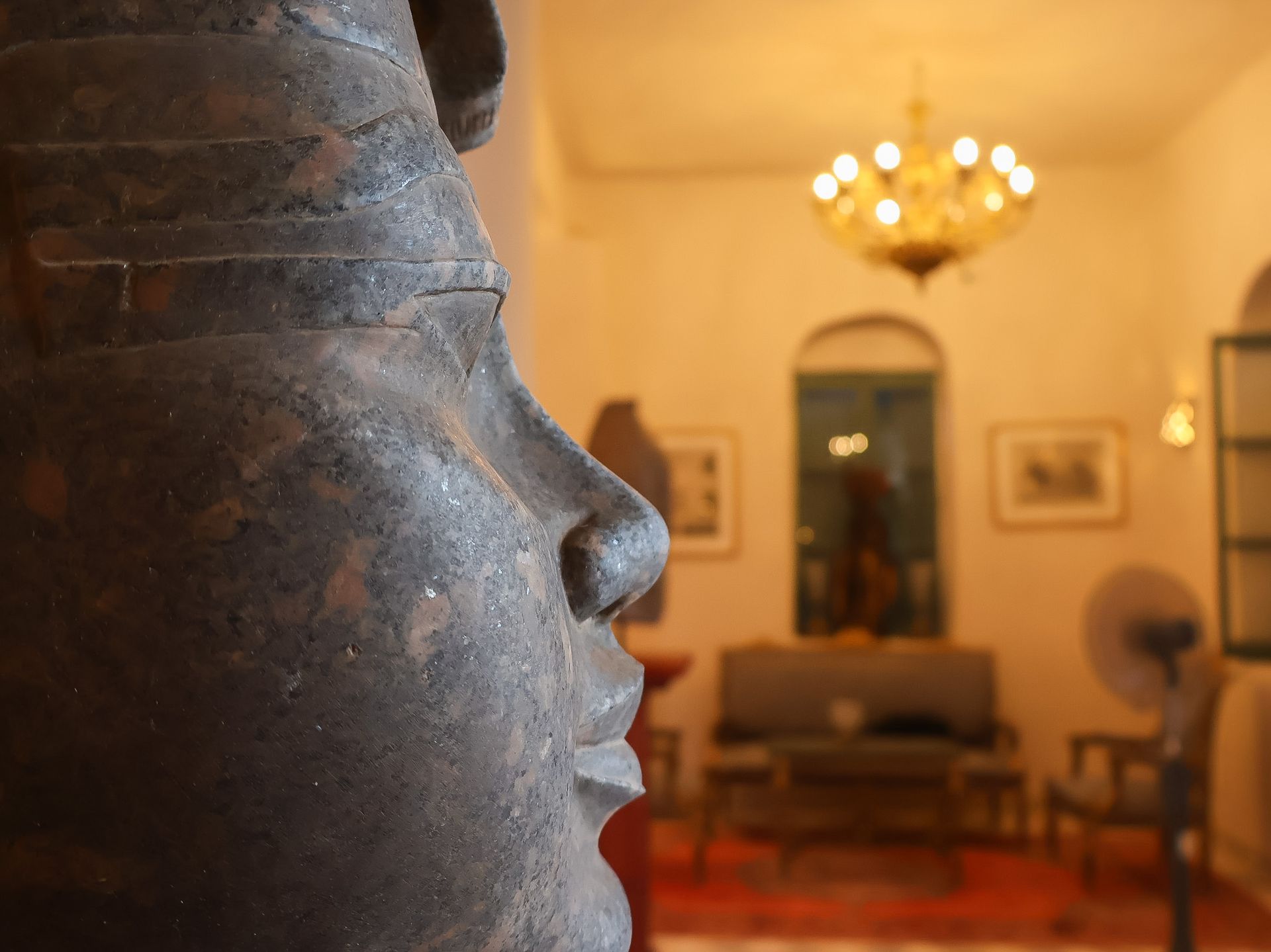 Close-up of a weathered stone face statue with a blurred room interior in the background.