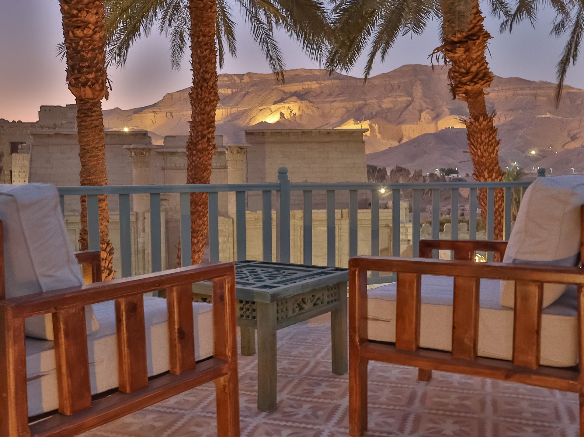Outdoor seating area overlooking ancient ruins and mountains, lit by warm evening light.