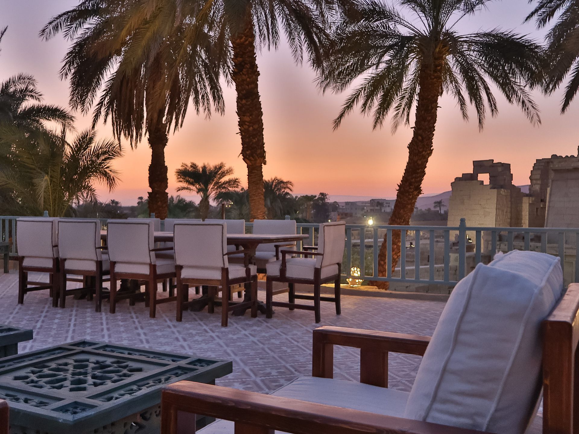 Rooftop terrace with seating, palm trees, and sunset view over a distant town.