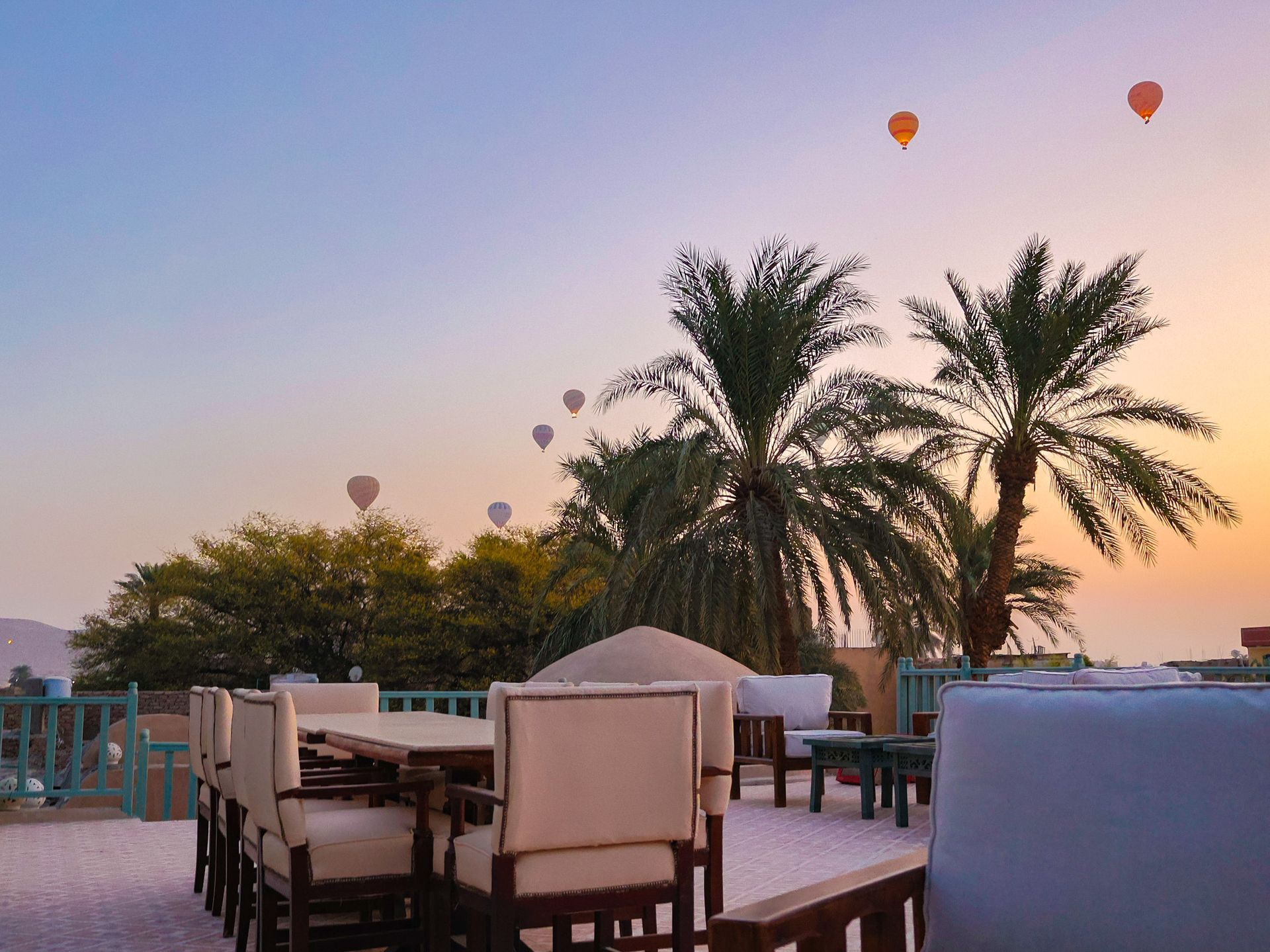 Outdoor dining area at sunset with palm trees and hot air balloons in the sky.