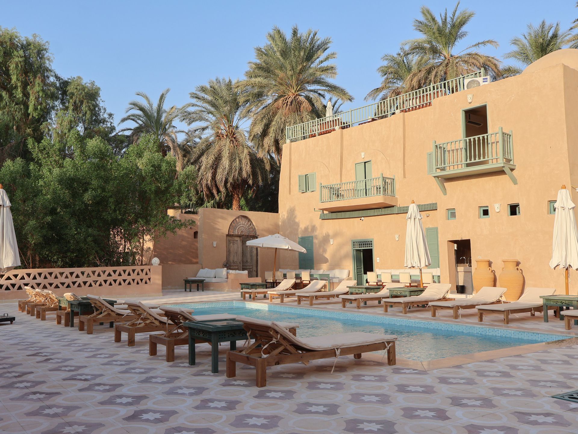 Poolside resort with sun loungers, clay-colored building, and palm trees.