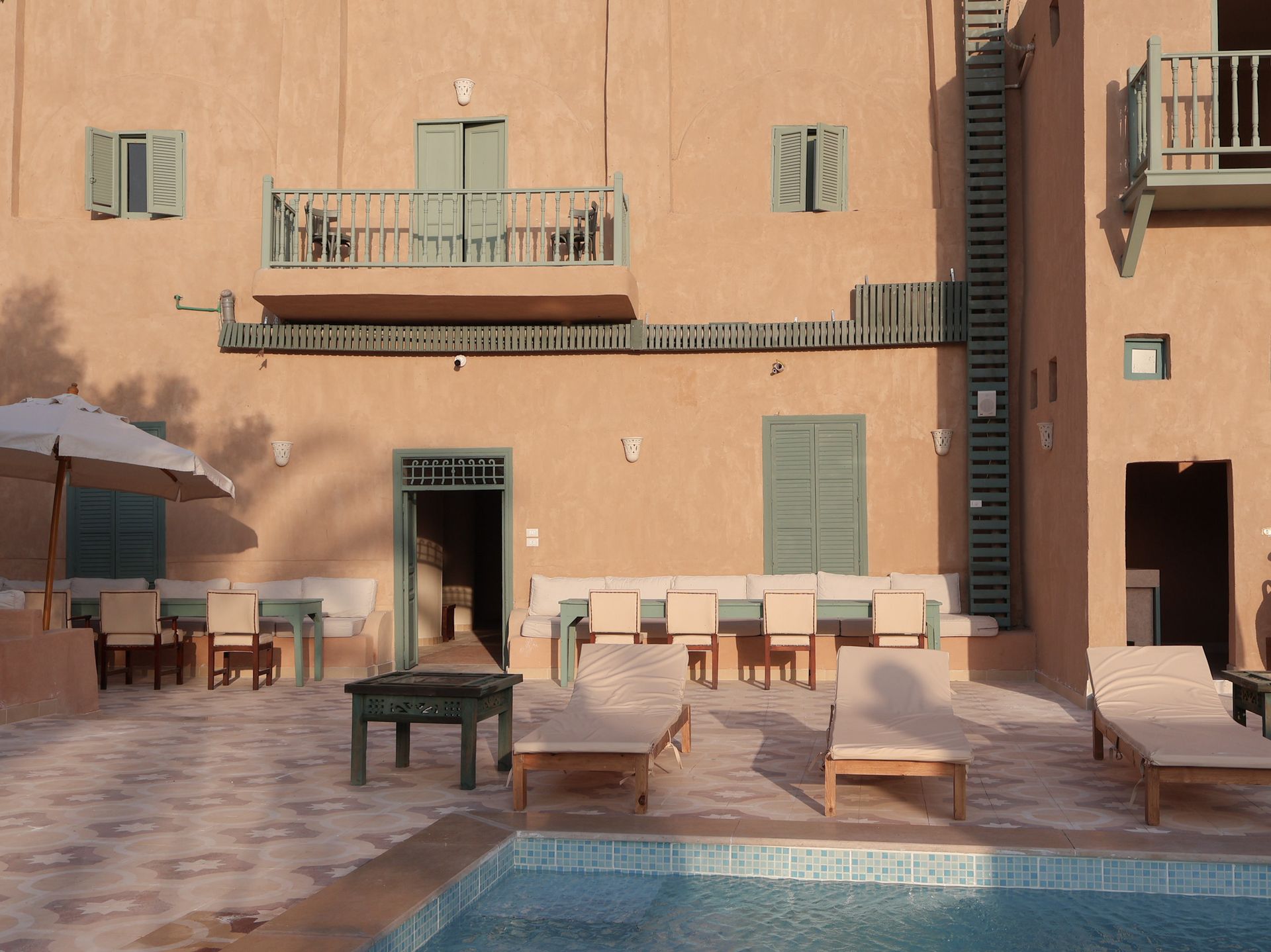 Courtyard with pool, beige building, teal shutters, balcony, and lounge chairs.