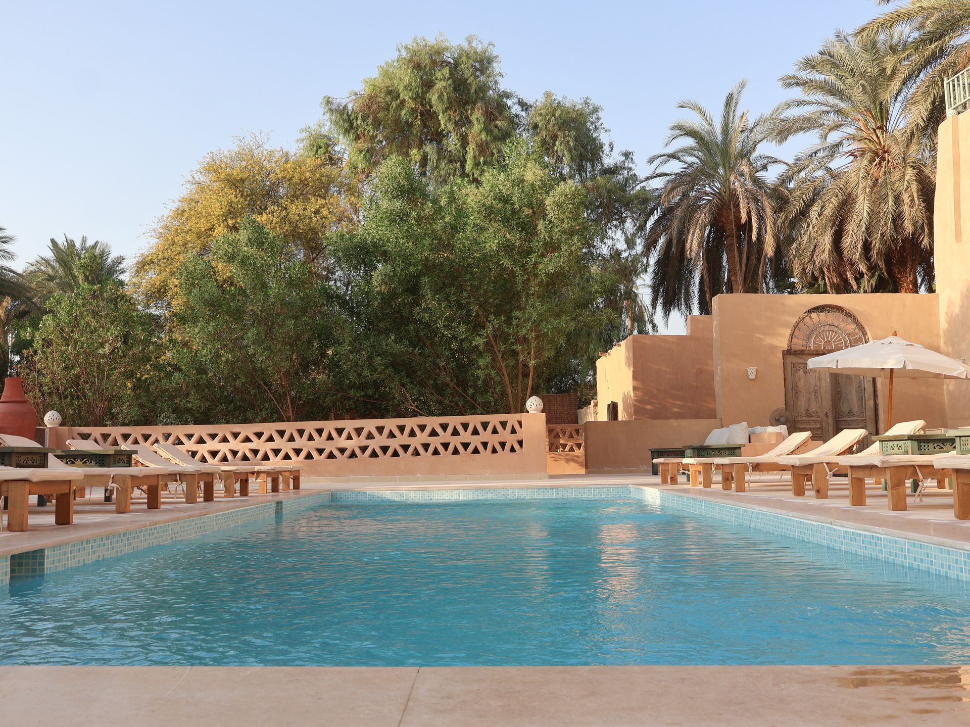 Swimming pool surrounded by sunbeds, trees, and buildings with a desert-like setting.