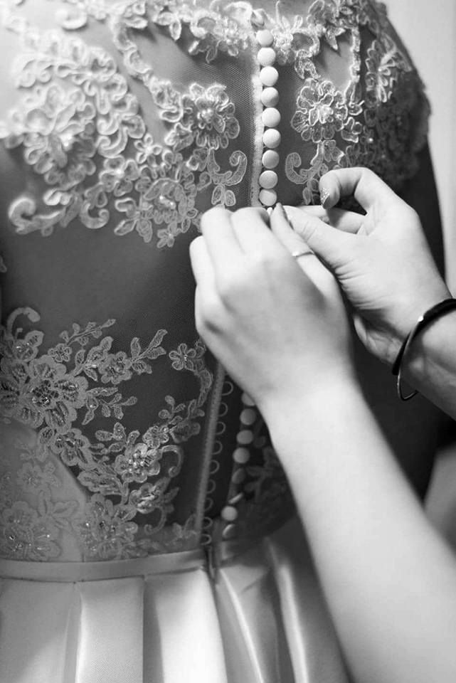 Hands buttoning the back of a white wedding dress, showcasing delicate lace detailing.