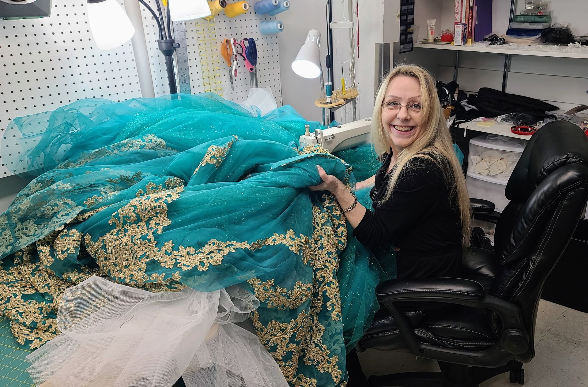 Woman sewing turquoise fabric with gold embroidery at a table with a sewing machine.