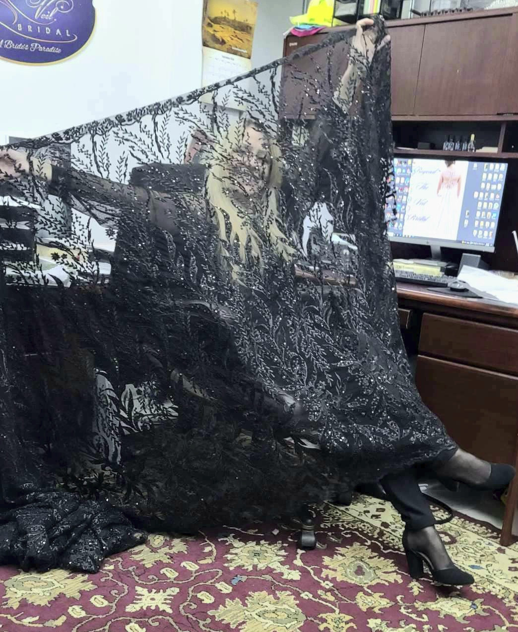Woman smiles, holding up a large, dark, sequined fabric in an office.