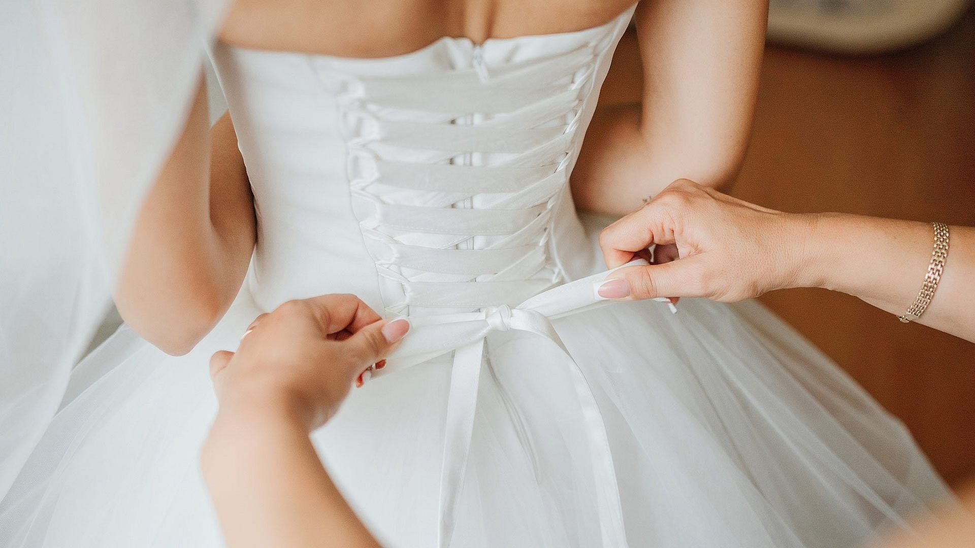 Bride in white dress, back view, being laced up by hands, preparing for wedding.