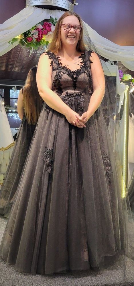Woman in a dark formal gown, smiling, standing indoors near other dresses.