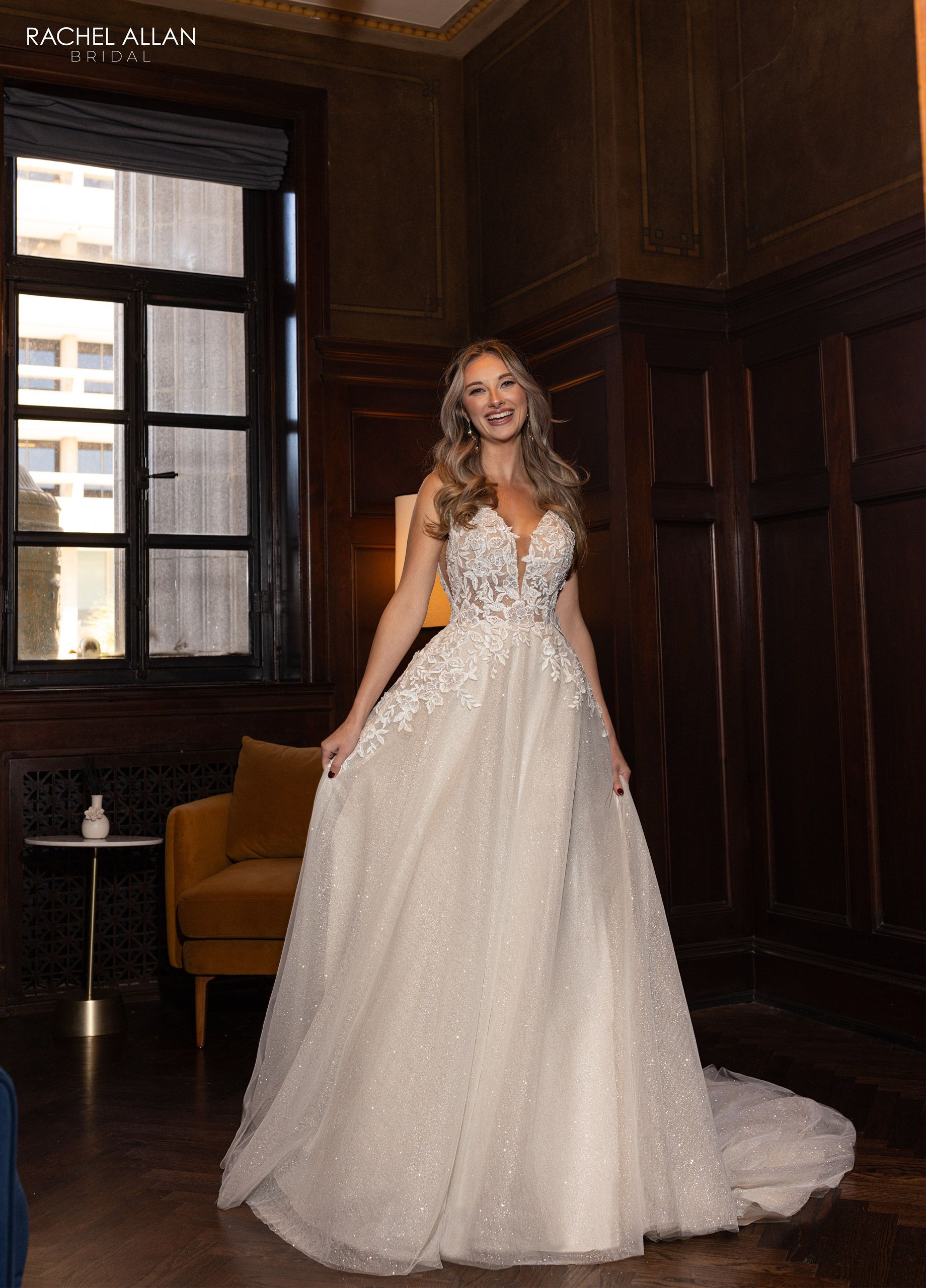 Woman in sparkling white wedding gown smiles in a room with wood paneling and a window.
