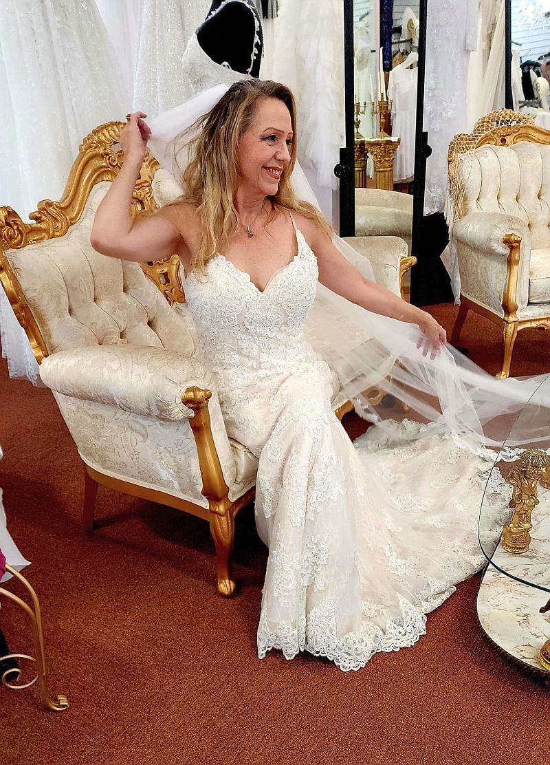 Woman in wedding dress, seated on ornate chair, holding veil in bridal shop.