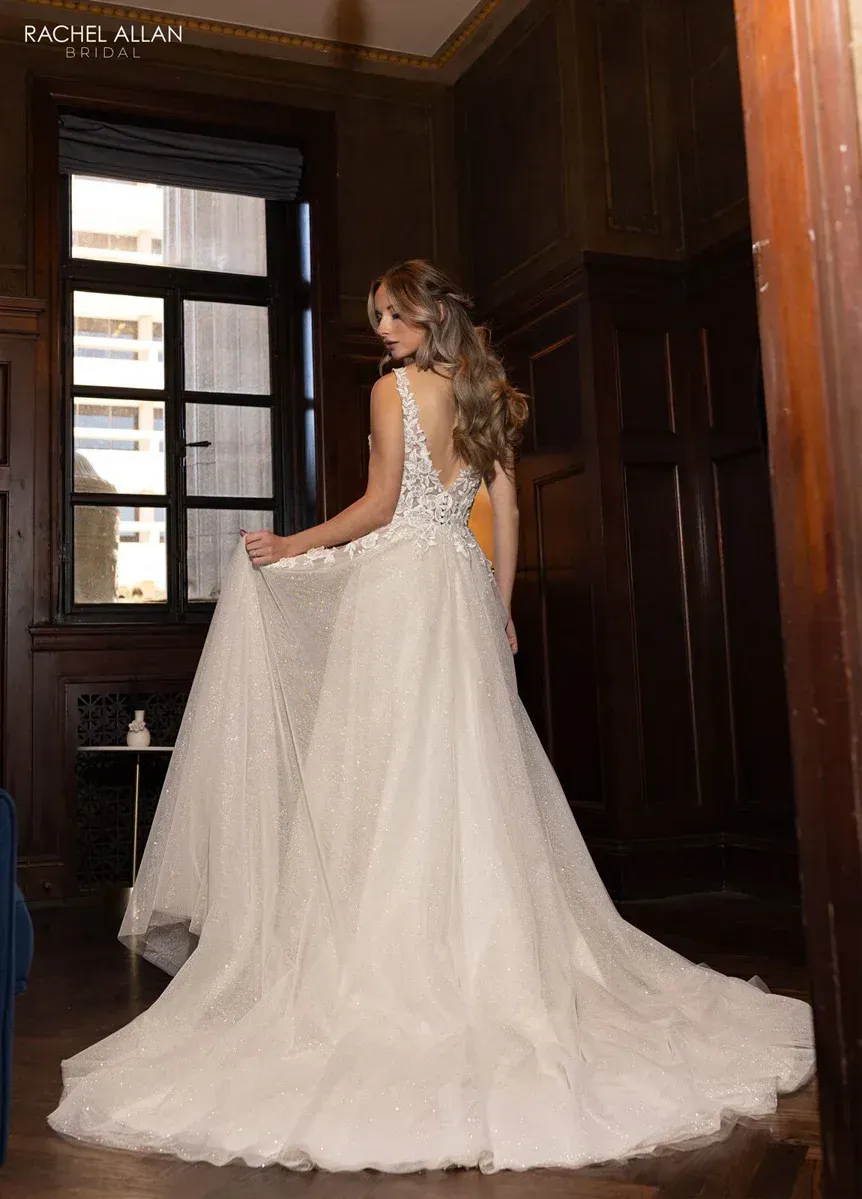 Woman in sparkling white wedding gown smiles in a room with wood paneling and a window.