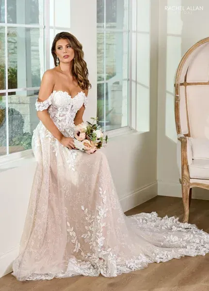 Woman in a white and blush off-the-shoulder wedding dress, posed in a room with a decorative chair.