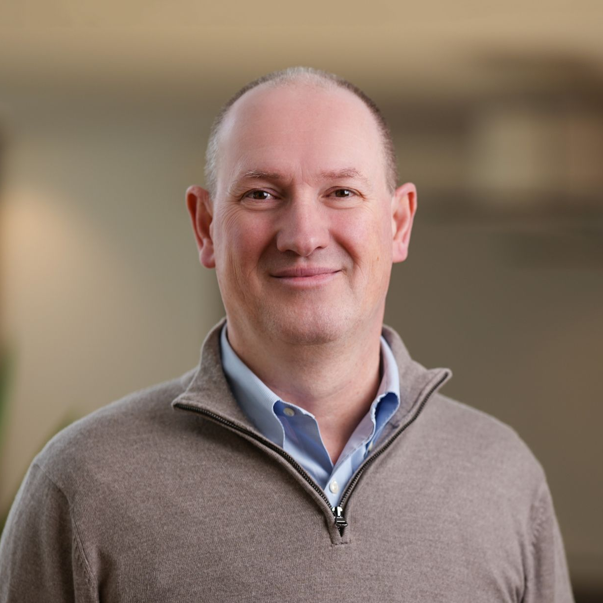 Smiling bald man in a gray sweater over a blue shirt against a softly blurred indoor background