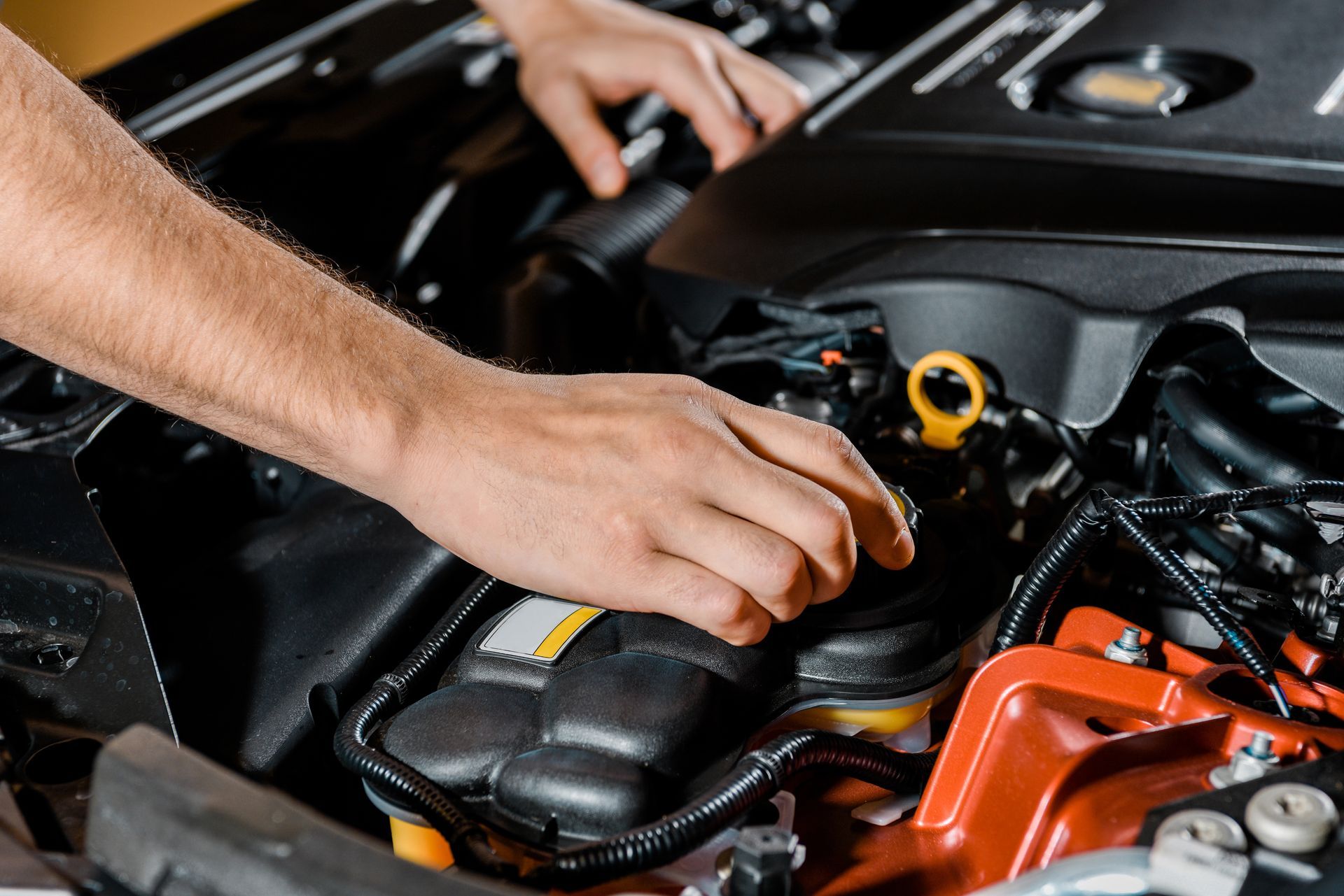 Mani che controllano l'olio motore nel vano motore nero e aperto di un'auto, con parti del motore arancioni visibili.