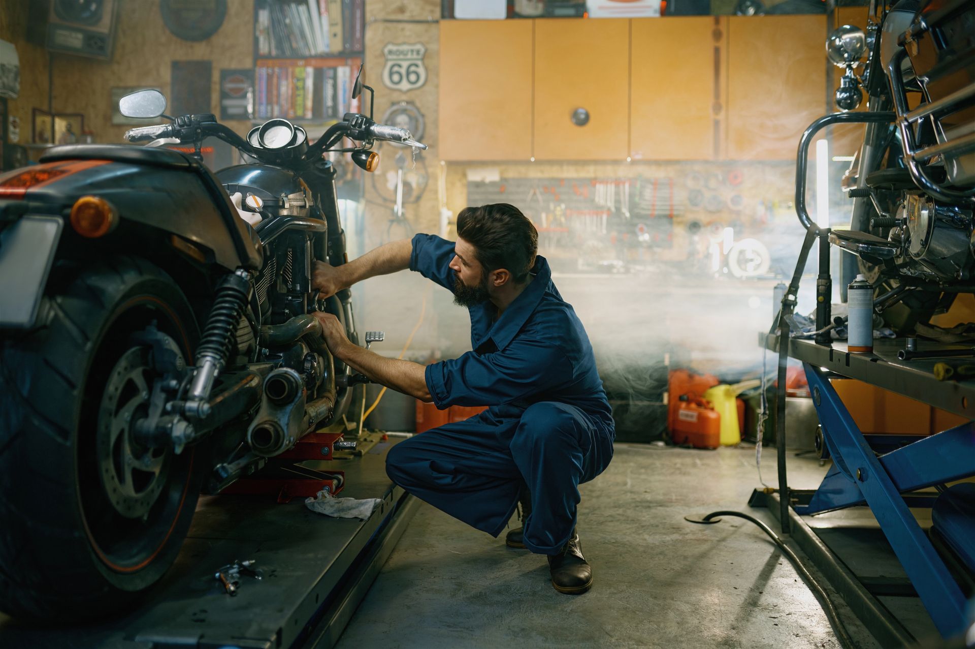 Un meccanico è accovacciato accanto a una motocicletta in un'officina, intento a utilizzare un attrezzo sotto una luce calda.