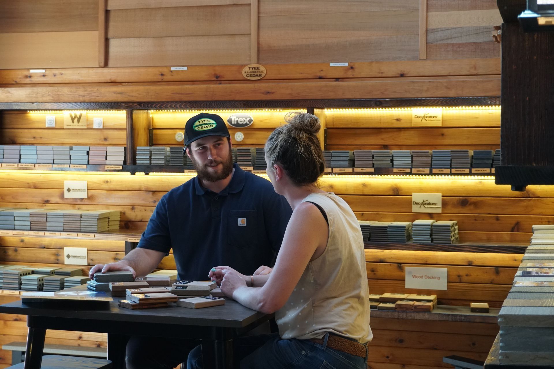 A man and woman at a table in a wood store, discussing samples. Shelves of wood behind them.