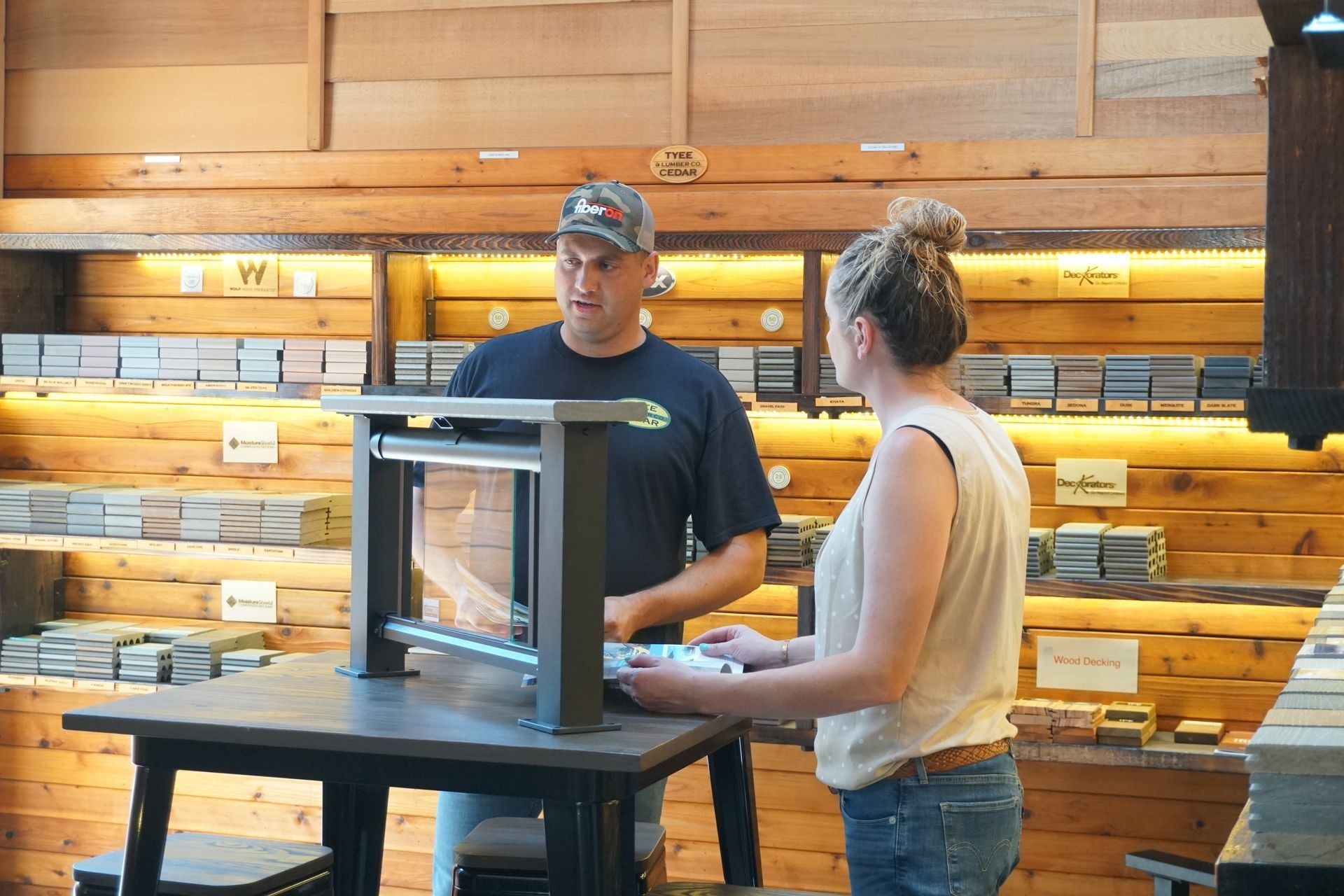 Man and woman examining a display table in a wood store, shelves of wood in background.