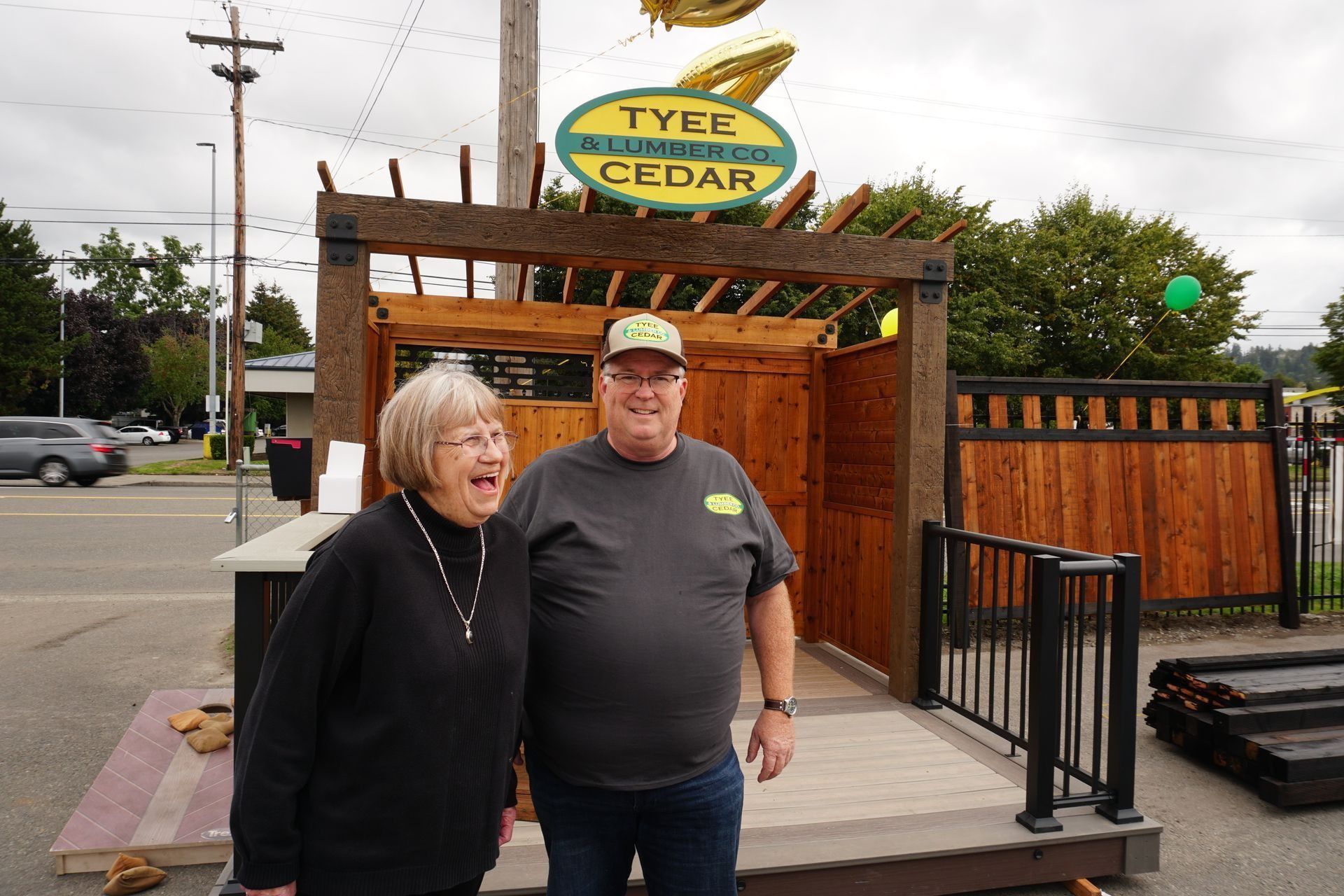 Two people stand in front of a wood structure with a sign that reads