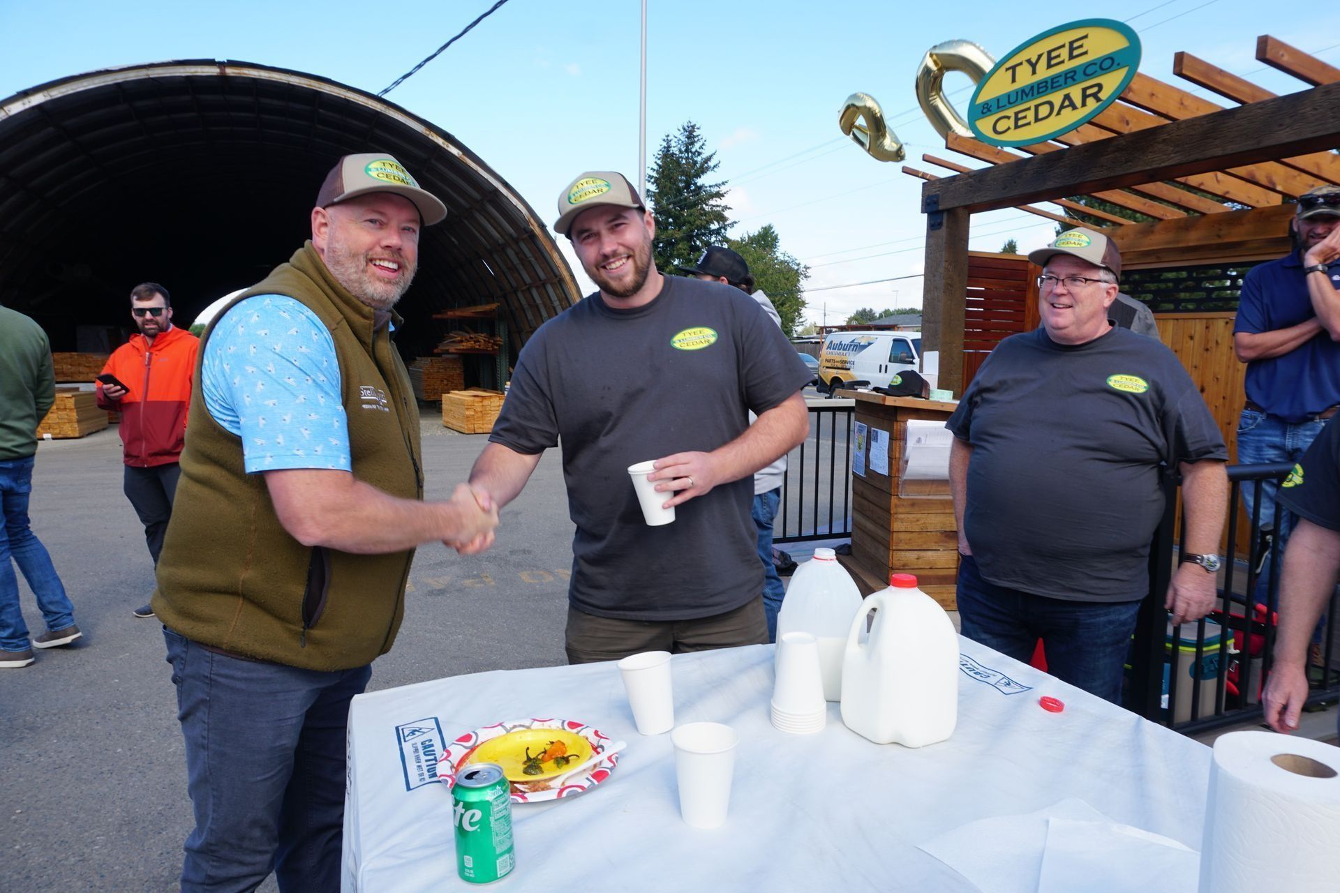 Three men shake hands at an outdoor event near a sign for