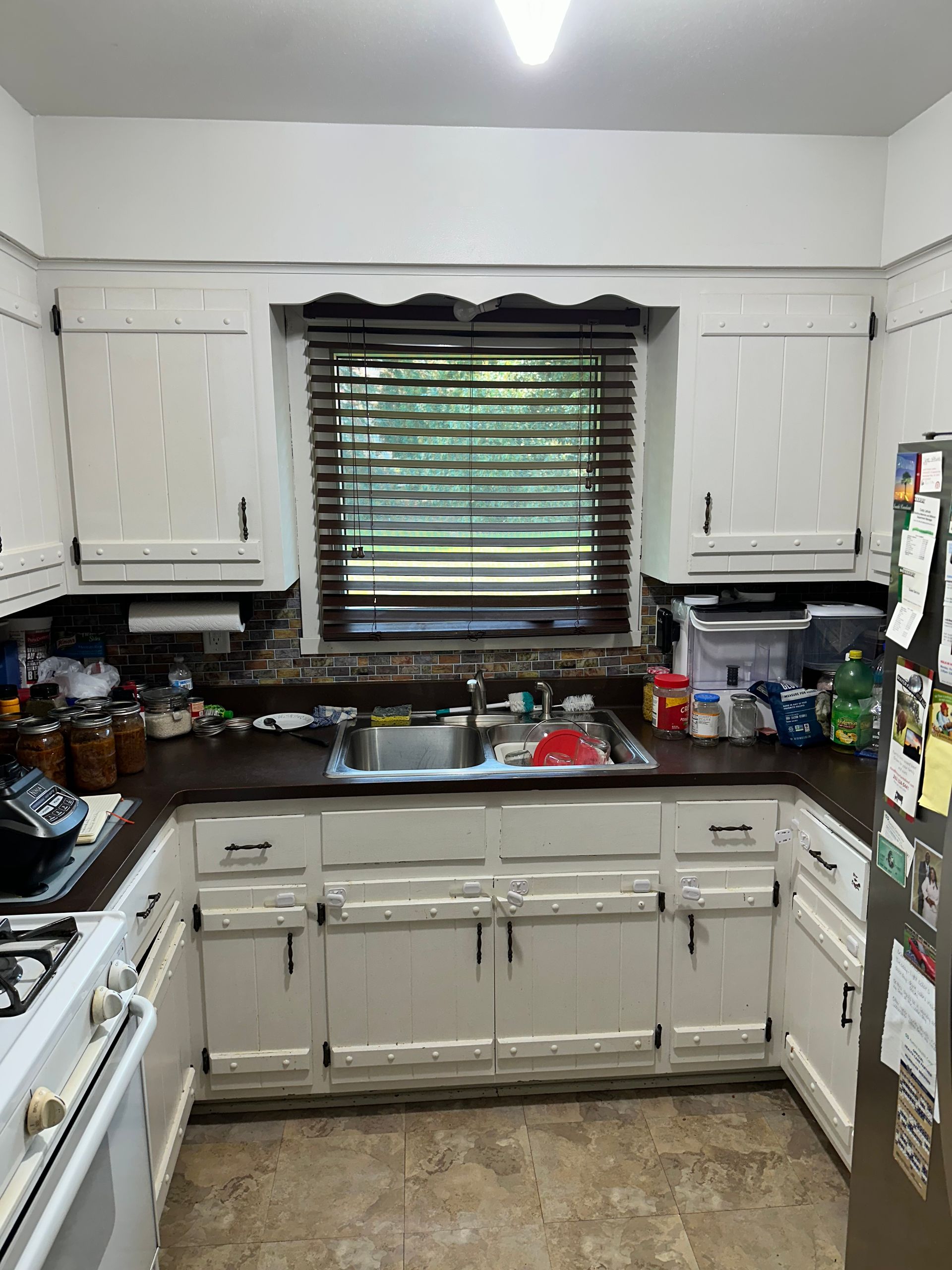 Cluttered white kitchen with sink under window, dark countertops, and stove at left