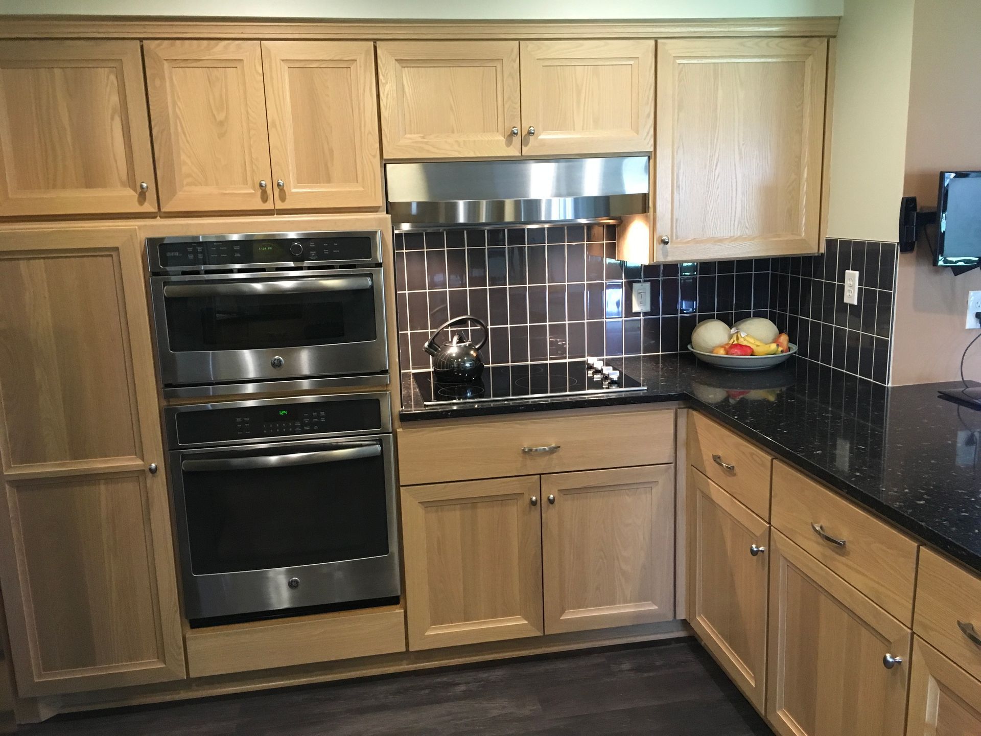 Modern kitchen with light wood cabinets, black countertops, stainless steel oven, and tiled backsplash.