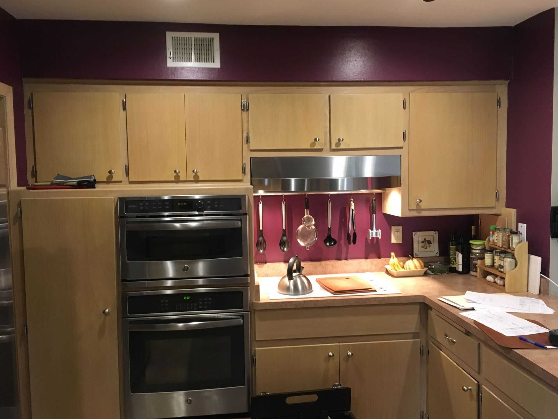 Kitchen with light wood cabinets, dark double oven, and countertop clutter under a stainless range hood.
