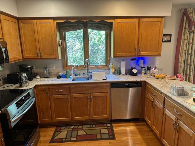 Kitchen with wood cabinets, double sink under window, dishwasher, oven, and patterned rug on the floor.