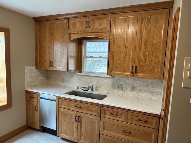 Wooden kitchen cabinets and sink area with stainless dishwasher, light countertops, and a window above the sink