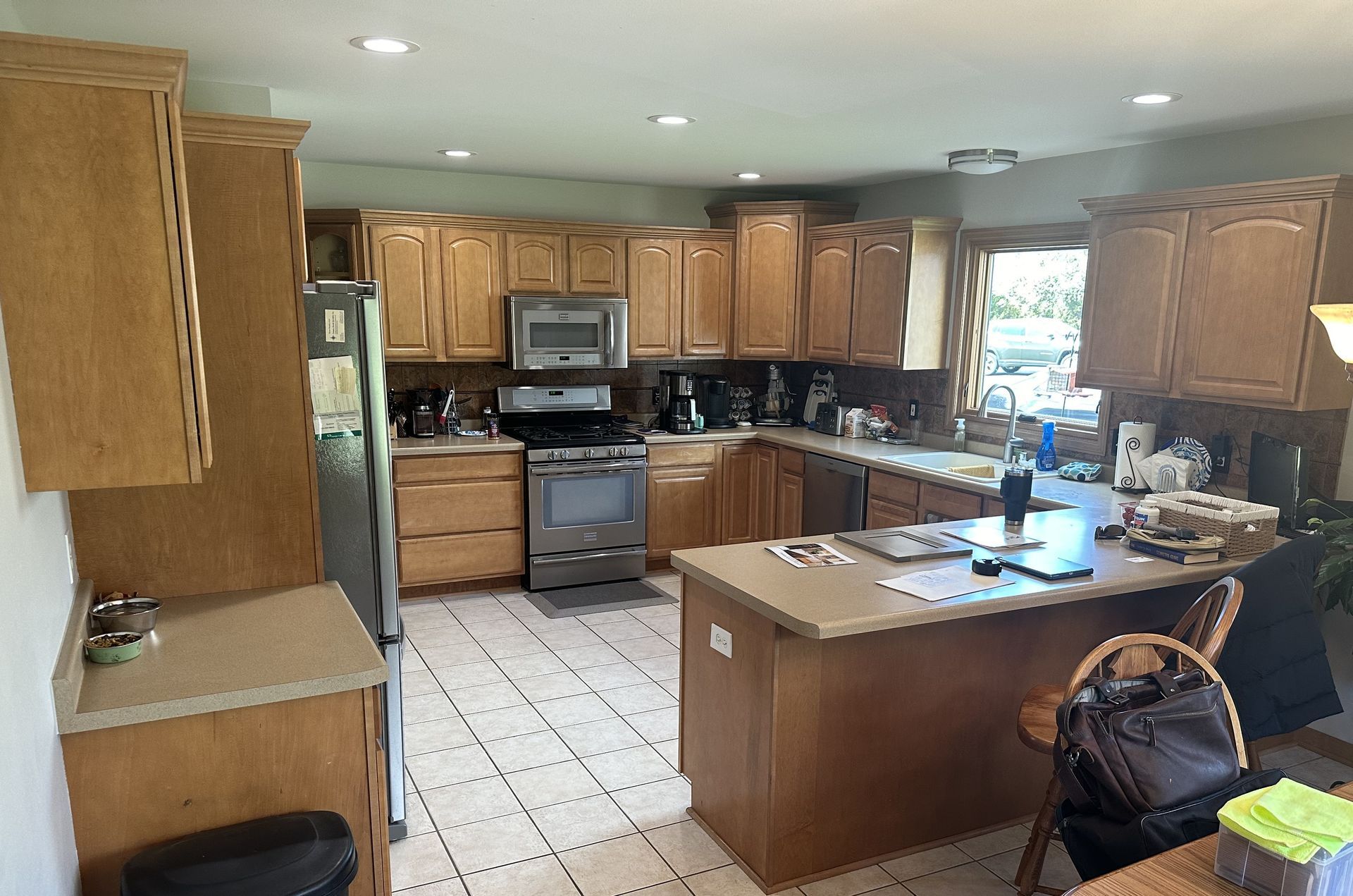 Bright kitchen with wood cabinets, white tile floor, black appliances, and a central island by a window.