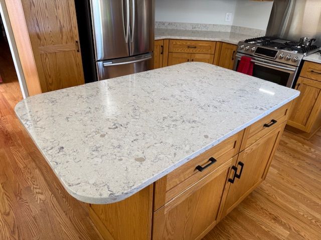 Kitchen island with white speckled countertop and wood cabinets, beside a stainless steel refrigerator and stove.