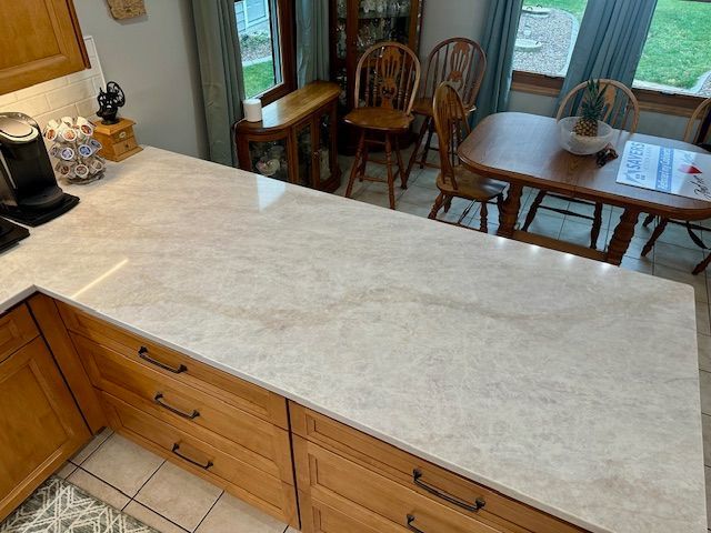 Kitchen island with a light stone countertop and wood cabinets, overlooking a dining area by large windows.