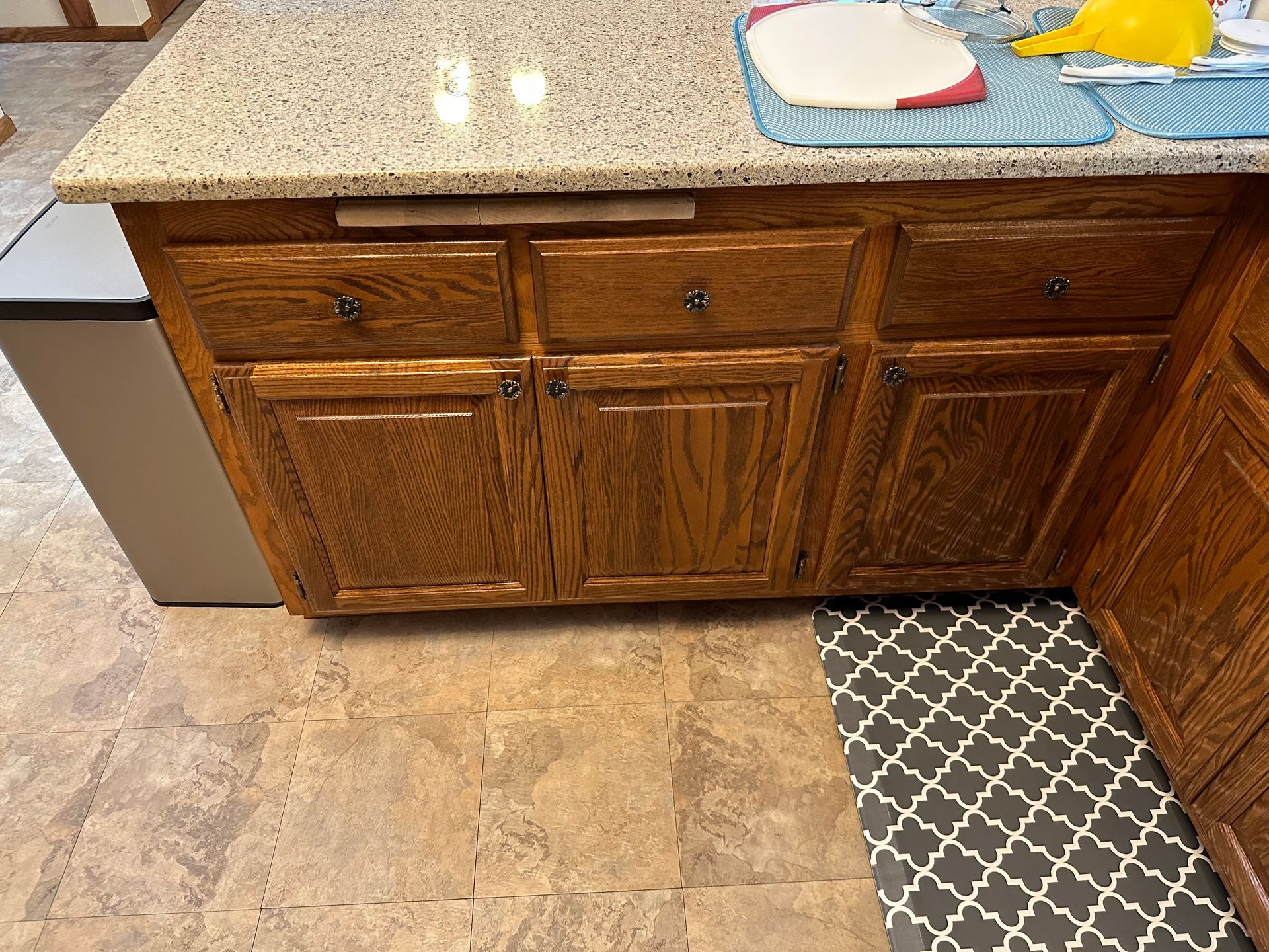 Kitchen cabinets below a speckled countertop with a white bowl and yellow sponge on top.