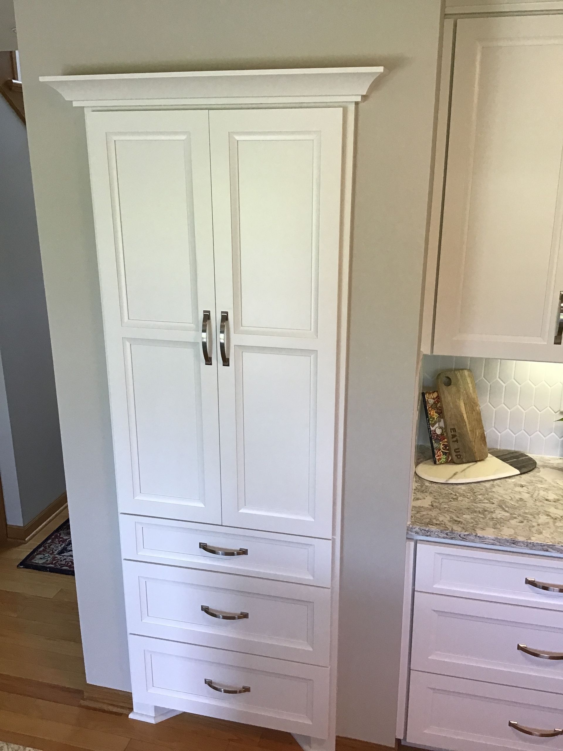 Tall white kitchen pantry cabinet with double doors, drawers, and black handles beside a countertop