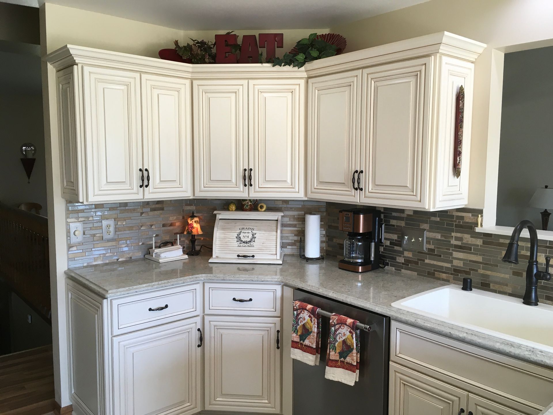 White kitchen with wood cabinets, tiled backsplash, coffee maker, and decorative “EAT” sign above the cabinets